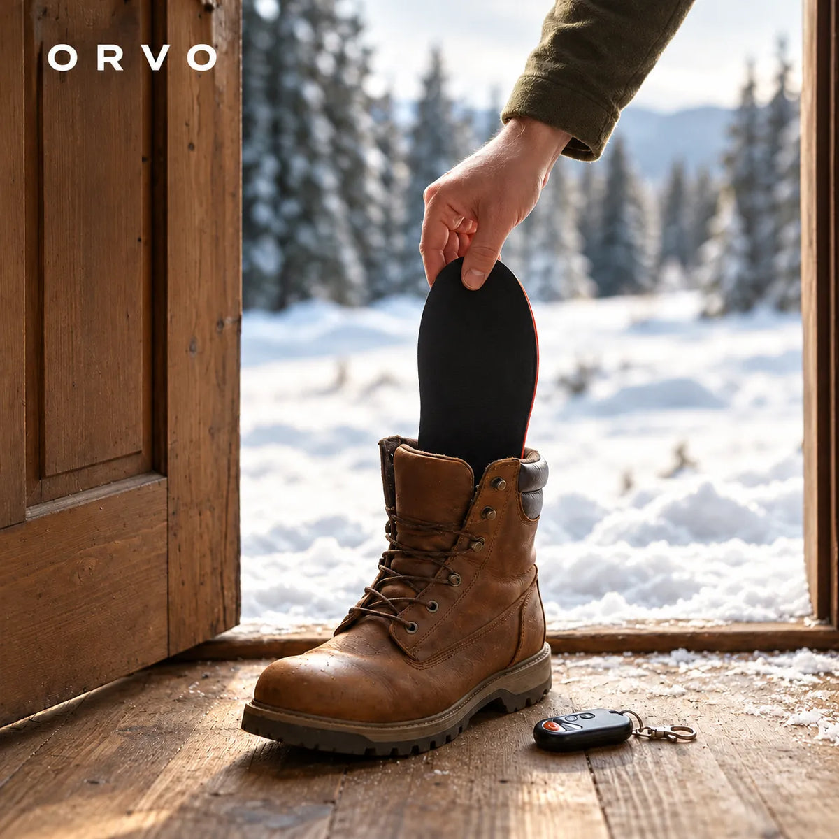 Hand lägger en värmesula i en känga vid en öppen dörr mot snöig skog, fjärrkontrollen på golvet